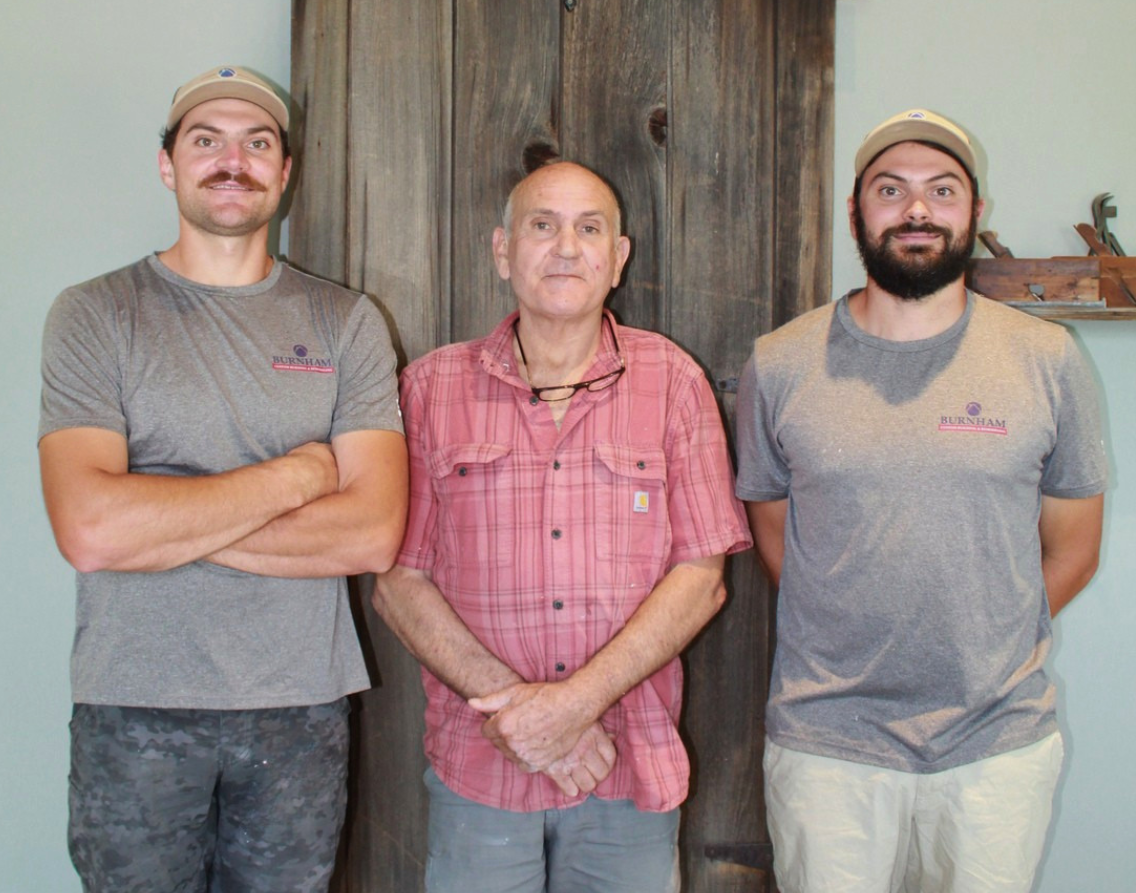 Paul, Ross, and Eli Burnham of Burnham Custom Building and Remodeling standing together in front of a reclaimed wood accent door